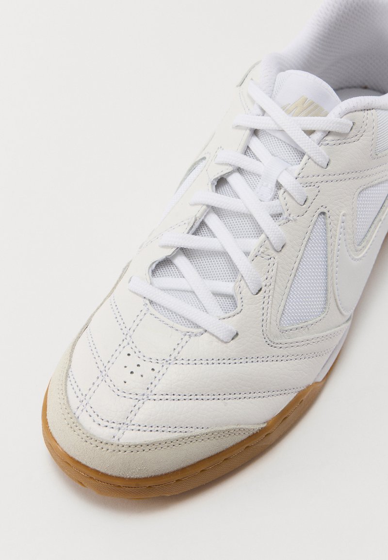 Zapatilla deportiva blanca con un empeine de cuero texturizado y malla, que presenta cordones blancos, suela de goma y perforaciones notables para la transpirabilidad.