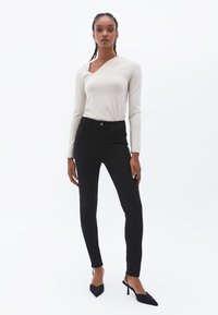 Blusa de manga larga color crema con un escote asimétrico, combinada con jeans negros ajustados y tacones puntiagudos negros con un sutil detalle de correa.