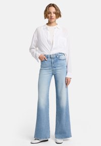 Jean en denim bleu clair évasé avec une taille haute, présentant un délavage usé et des détails de froissage subtils, associé à une chemise blanche à boutons.