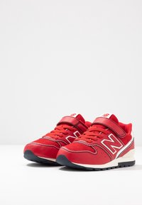 Une paire de baskets rouges New Balance pour enfants avec des lacets, une sangle Velcro, des semelles intermédiaires blanches et des semelles extérieures noires sur une surface blanche.