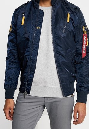 Homme portant une veste bomber bleu marine avec des tirettes de fermeture éclair jaunes, un pull gris et un pantalon gris, debout les bras détendus le long du corps.