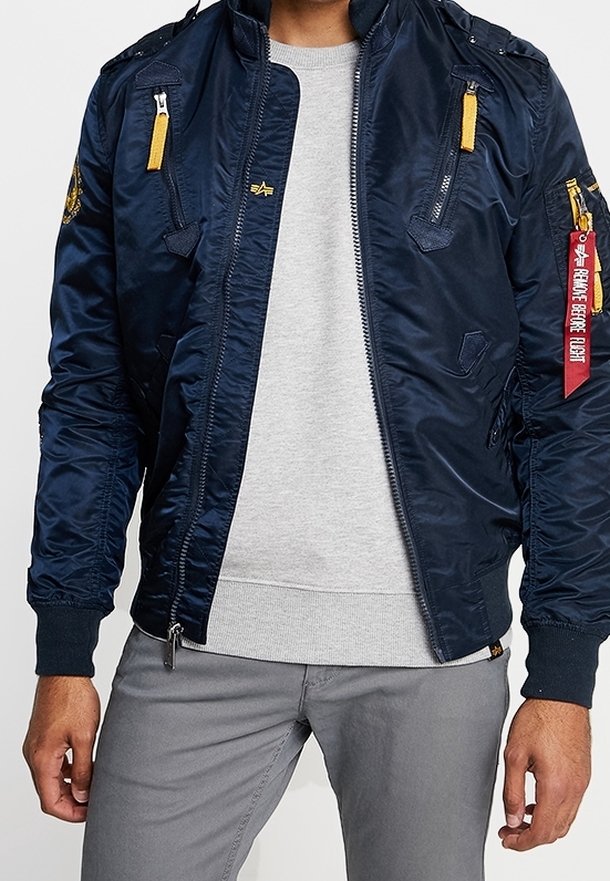 Homme portant une veste bomber bleu marine avec des tirettes de fermeture éclair jaunes, un pull gris et un pantalon gris, debout les bras détendus le long du corps.