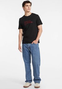 T-shirt noire à manches courtes avec l'imprimé rouge "GUESS JEANS", associée à un jean en denim bleu clair et des baskets beiges avec des accents blancs.