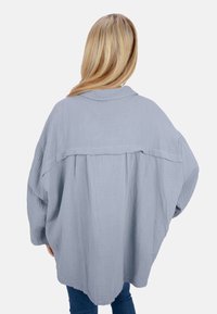 Oversized light blue shirt jacket made of textured fabric, featuring a back yoke detail, wide sleeves, and a rounded hem.