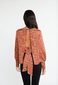Achteraanzicht van een rood en beige geblokte blouse met lange mouwen, voorzien van een sleufopening en een striksluiting op de rug, gemaakt van zachte stof.