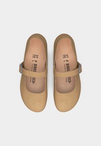 Τακούνια Birkenstock σε καφέ ανοιχτό χρώμα με στρογγυλεμένα δάχτυλα και μονό λουρί που κλείνει με ασημένιες πόρπες, εμφανιζόμενα από πάνω σε λευκό φόντο.