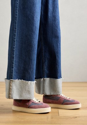 Różowe i szare zamszowe sneakersy z kremowymi sznurówkami, zestawione z szerokimi ciemnoniebieskimi dżinsami z postrzępionym jasnym denimu u dołu.