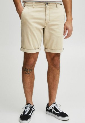 Beige cotton shorts with rolled cuffs, featuring front pockets and a button closure, paired with black sneakers and white shoelaces.