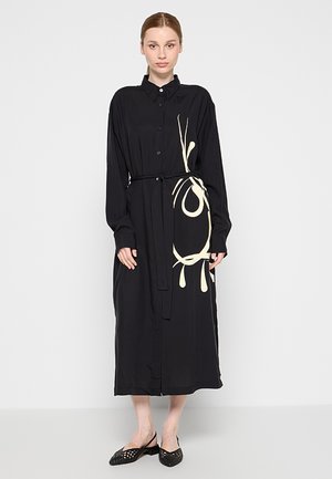 Kobieta nosząca długą czarną sukienkę z guzikami z beżowym abstrakcyjnym wzorem po boku, przewiązaną w talii oraz czarne plecione buty typu slingback.