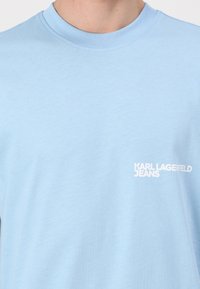 T-shirt en coton bleu clair avec un col rond et une texture douce. Porte un logo blanc "KARL LAGERFELD JEANS" sur le côté gauche.