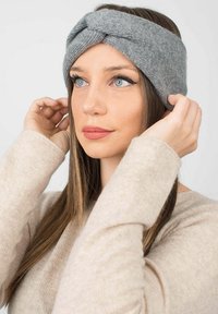 Fascia grigia lavorata a maglia con un design attorcigliato, texture morbida e larghezza media. Indossata da una persona con capelli lisci e un maglione chiaro.