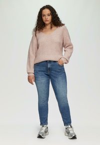 Suéter de punto acanalado de color rosa claro con escote en V, combinado con jeans de mezclilla azul y zapatillas plateadas. Ajuste y diseño casual y cómodo.