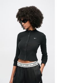 Μαύρο ριμπ μακρυμάνικο τοπ με γιακά με κουμπιά και λευκό λογότυπο της Nike. Φορώντας το με μαύρο σατέν παντελόνι που διαθέτει λαστιχένια μέση με το λογότυπο της εταιρείας.