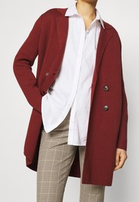 Burgundy dubbelknäppt kappa med stora knappar, ribbad textur och sjalkrage, bärs över en vit skjorta och byxor med houndstooth-mönster.