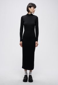 Crna haljina s dugim rukavima i visokim ovratnikom, pripijena silueta s bočnim prorezima, izrađena od glatkog, rastezljivog materijala.