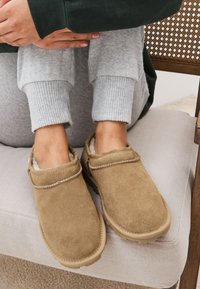 Mocassini in camoscio marrone con fodera interna morbida, suole piatte e cuciture decorative. Indossati con pantaloni della tuta grigi a coste.