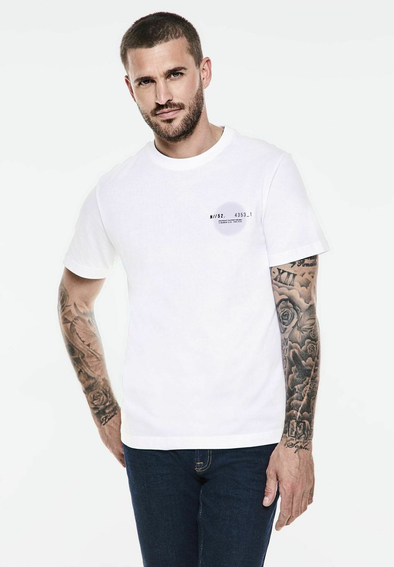 Bílý krátký rukáv tričko s kulatým výstřihem, na levé straně s malou grafickou nálepkou. Měkká bavlněná tkanina s hladkou texturou.