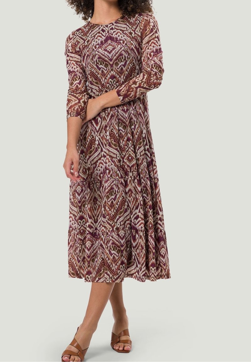 Robe longue avec motif abstrait en bordeaux, crème et marron. Manches longues, encolure ronde, et jupe plissée avec une coupe décontractée.