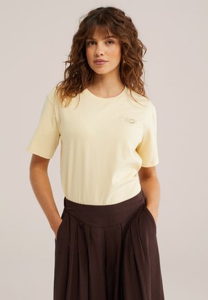 Donna con capelli ricci castani che indossa una maglietta a maniche corte di colore giallo pallido e una gonna plissettata a vita alta di colore marrone scuro, mani nelle tasche, sfondo neutro.