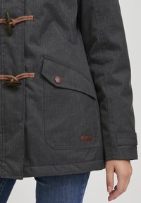 Mörkgrå jacka med toggleknappar, ficka med klaff och tryckknapp, läderlogotyp, bärs med blå jeans och en synlig högerhand.