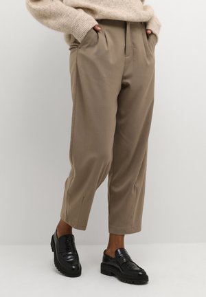 Personne portant un pantalon beige taille haute à jambes larges avec les mains dans les poches et des mocassins en cuir noir épais, debout devant un fond uni.