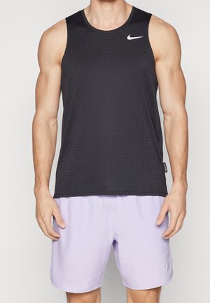 Hombre con camiseta deportiva negra sin mangas de Nike y pantalones cortos de color púrpura claro, de pie frente a un fondo claro y liso.