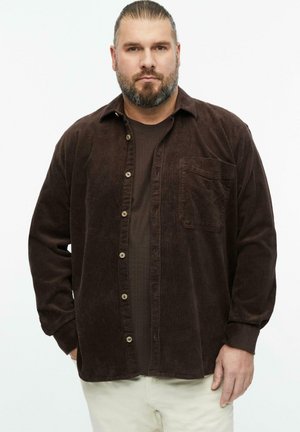 Mann mit Bart und nach hinten gekämmtem Haar, der ein dunkelbraunes Cordhemd und beige Hose trägt und mit den Händen in den Taschen steht.