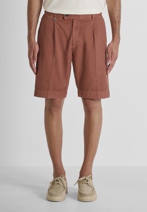 Hombre con pantalones cortos plisados de color óxido y zapatos beige con cordones, de pie sobre un fondo liso de color gris claro.