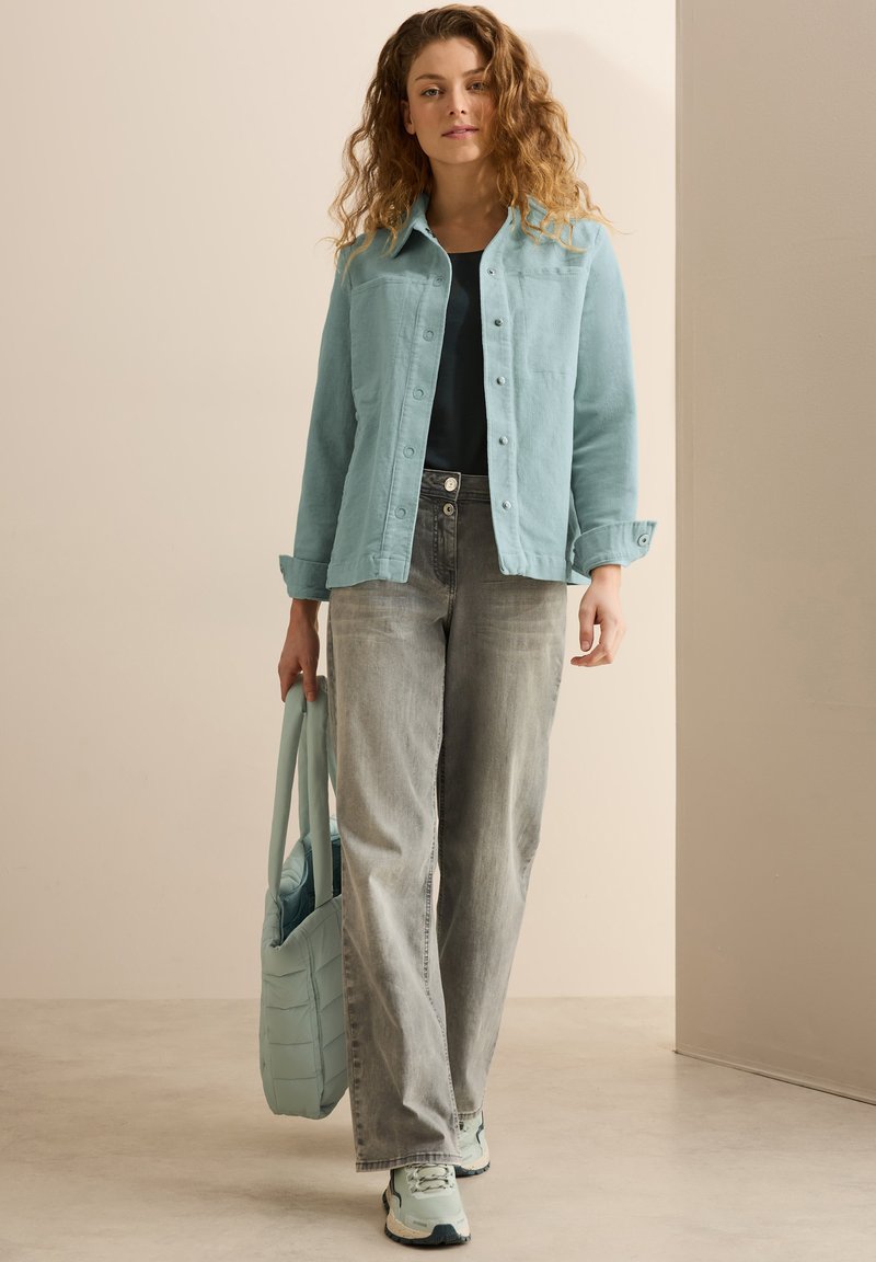 Giacca di denim azzurro chiaro con bottoni, top nero, jeans grigi e sneakers azzurro chiaro. Porta una borsa a tracolla trapuntata azzurro chiaro.