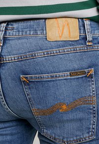Jeans de mezclilla azules con un parche de cuero marrón claro que presenta un logotipo en relieve. Costuras naranjas visibles y un detalle ondulado único en los bolsillos traseros.