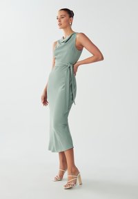 Sleek, sleeveless, mint green dress with a cowl neckline, waist tie, and flared hem, paired with white strappy high-heeled sandals.