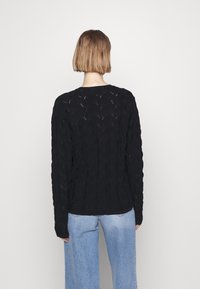 Persona con capelli corti castano chiaro che indossa un maglione nero di tessuto e jeans azzurri chiari, in piedi di fronte a uno sfondo bianco.