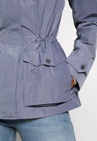 Gros plan d'une personne portant une veste Barbour violet clair avec des poches à rabat boutonnées et la main dans la poche latérale, associée à un jean bleu.