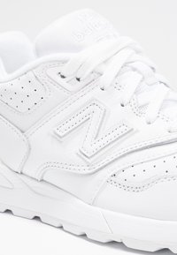 Bílé sportovní tenisky z kůže s perforovanými detaily, texturovaným logem N a plochými bílými šňůrkami. Tlustá bílá podrážka.