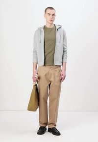 Felpa grigia con zip sopra una t-shirt verde oliva, pantaloni beige e scarpe nere. Tiene una borsa beige. Sfondo neutro.