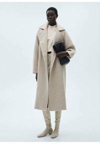 Beige Wollmantel, lang und oversized, mit Reverskragen. Kombiniert mit einem leichten Pullover und hochgeschnittenen Hosen, ergänzt durch kniehohe beige Stiefel.