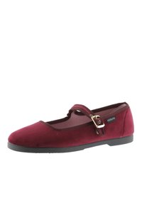 Zapato plano de terciopelo burdeos con punta redonda, correa al tobillo con hebilla dorada y suela de goma texturizada. Etiqueta con logotipo en el lateral.
