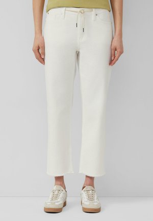 Person trägt cremefarbene, gekürzte Hosen mit ausgefransten Säumen und weiße Turnschuhe mit beigen Akzenten, steht vor einem hellgrauen Hintergrund.