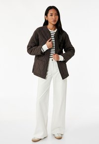 Nadto quiltowana brązowa kurtka z okrągłym dekoltem, o gładkiej fakturze i bocznymi kieszeniami, zestawiona z pasiastą koszulą i szerokimi, białymi spodniami.