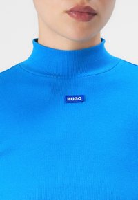 Moteris vilki ryškiai mėlyną raštuotą polo megztinį su maža etiketė "HUGO", esanti vos po kaklu.