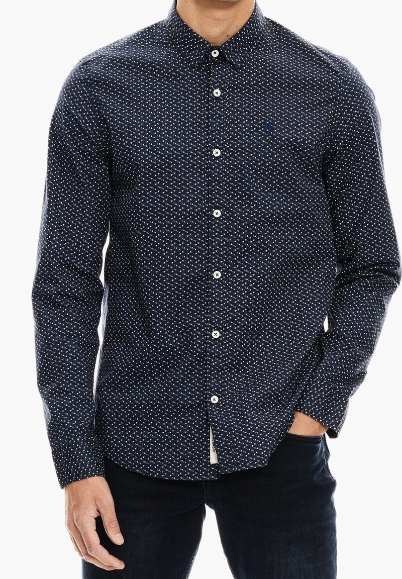Homme portant une chemise bleu marine à manches longues avec un petit motif blanc, boutonnée, avec une main dans la poche d'un pantalon foncé.