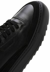 Baskets noires avec un dessus en cuir lisse et en daim, semelle en caoutchouc texturé, lacets plats et détails cousus sur les côtés.