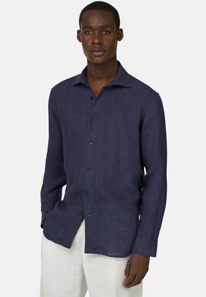 Homme portant une chemise à manches longues bleu marine boutonnée et un pantalon blanc, debout avec une expression neutre devant un fond uni.