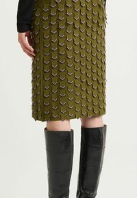 Jupe vert olive avec des écailles texturées et superposées, détails de couture violette, associée à des bottes noires élégantes montant jusqu'au genou, ornées d'un motif reptile.