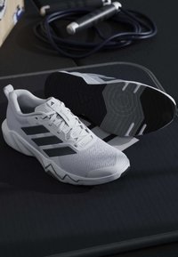 Λευκά αθλητικά παπούτσια Adidas με μαύρες ρίγες πάνω σε μαύρη επιφάνεια δίπλα σε μαύρο σχοινάκι για άλμα, τυλιγμένο σε ρολό, με γκρι χερούλια.