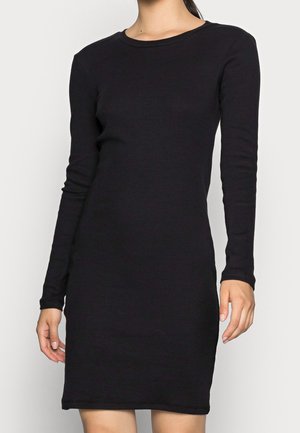 Kobieta ubrana w dopasowaną, czarną dzianinową sukienkę z długim rękawem i okrągłym dekoltem, stojąca na jasnym tle.