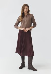 Brauner Strickcardigan über einem bordeauxfarbenen, plissierten Rock, kombiniert mit knielangen braunen Lederstiefeln. Der neutrale Hintergrund hebt die Details der Kleidung hervor.