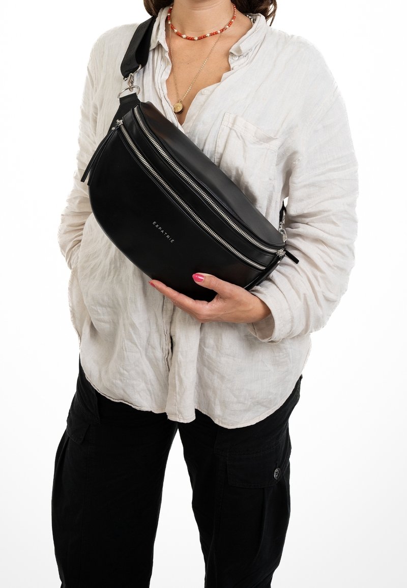 Personne portant une chemise blanche et un pantalon noir, tenant un sac bandoulière noir avec des fermetures éclair argentées portant l'inscription "EXPATRIÉ".