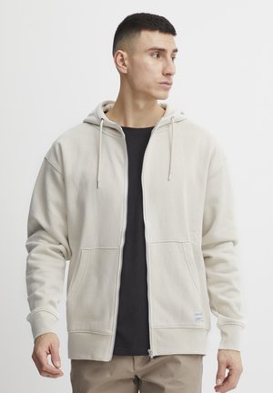 Sudadera con cremallera de color beige claro, con capucha ajustable y dos bolsillos frontales. Llevada sobre una camiseta negra. Textura de tela suave.