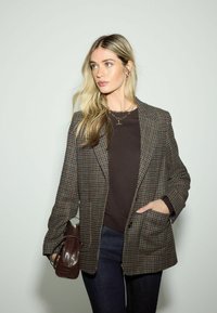 Blondynka ubrana w kratkowaną marynarkę, ciemną bluzkę, dżinsy, trzymająca brązową torebkę, patrząca lekko w lewo na tle jednolitego jasnego tła.